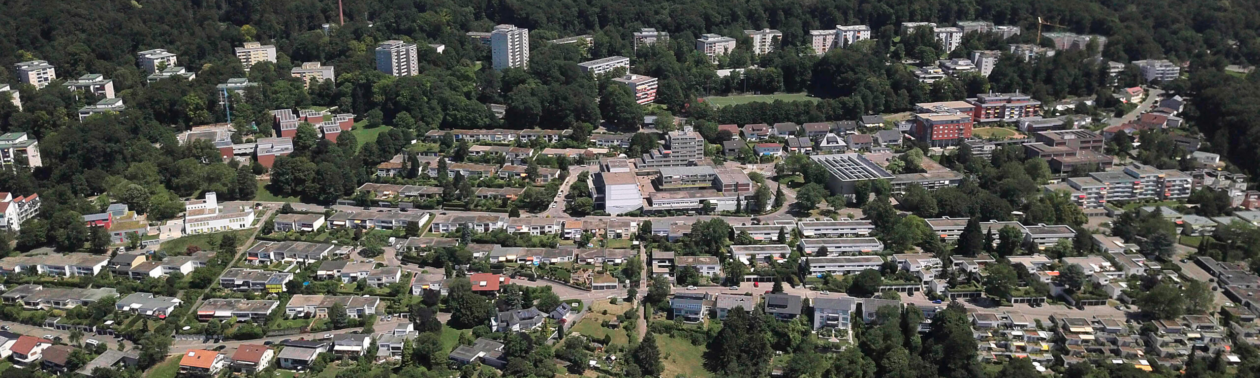 ein Drohnenbild vom gesamten Stadtteil, vom Westen her gesehen. Man erkennt die Hochhäuser am Waldrand und auf dem Feld davor die Ein- und Mehrfamilenhäuser